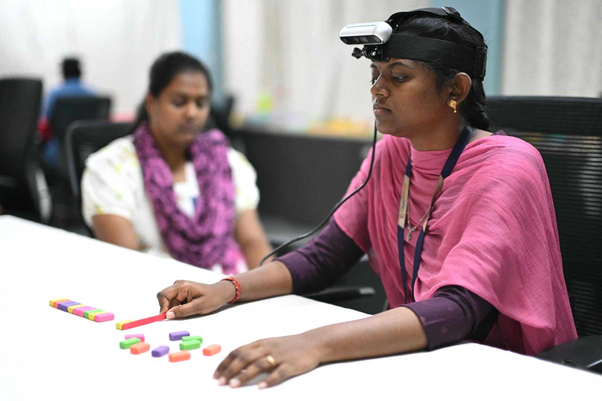 This photograph, taken on May 13, 2026, shows a worker (right) wearing an RGB camera on her head recording actions through motion capture while arranging coloured blocks at AI data company Objectways’ office in Tamil Nadu’s Karur district. Photo: AFP